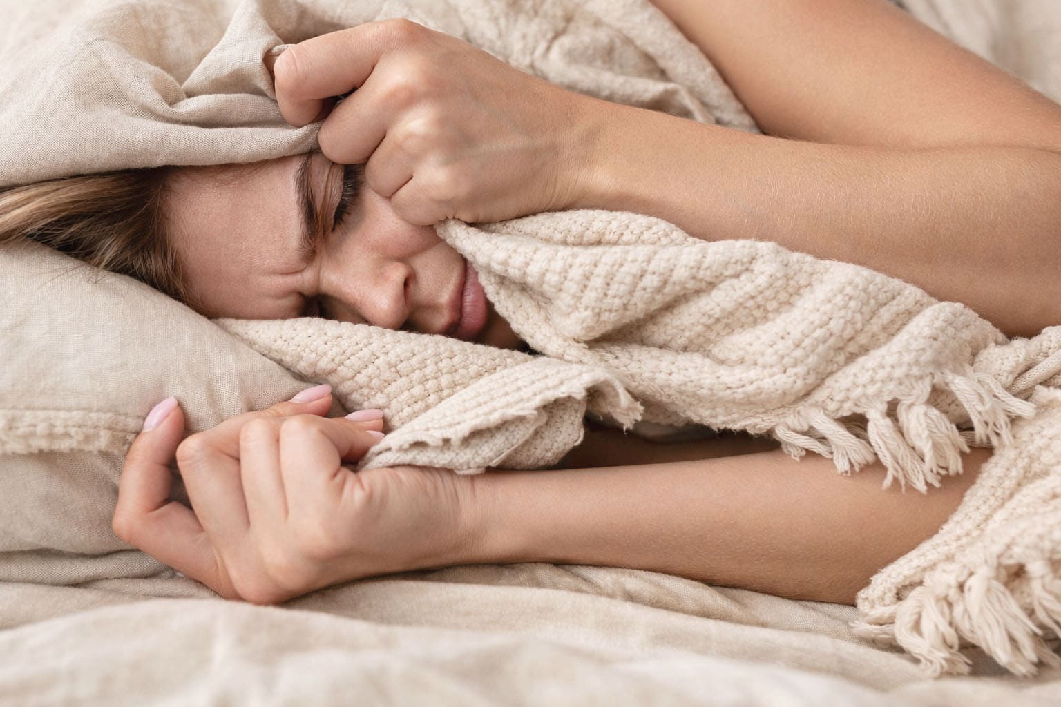 Femme allongée dans des draps beige et un plaid bohème crème, se cachant partiellement le visage dans la couette, ambiance cocooning et glamour, illustrant la fatigue pendant les règles.